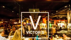 Un bar concurrido con mesas y clientes, un letrero visible que dice 'VICTOR'S', e iluminación interior.