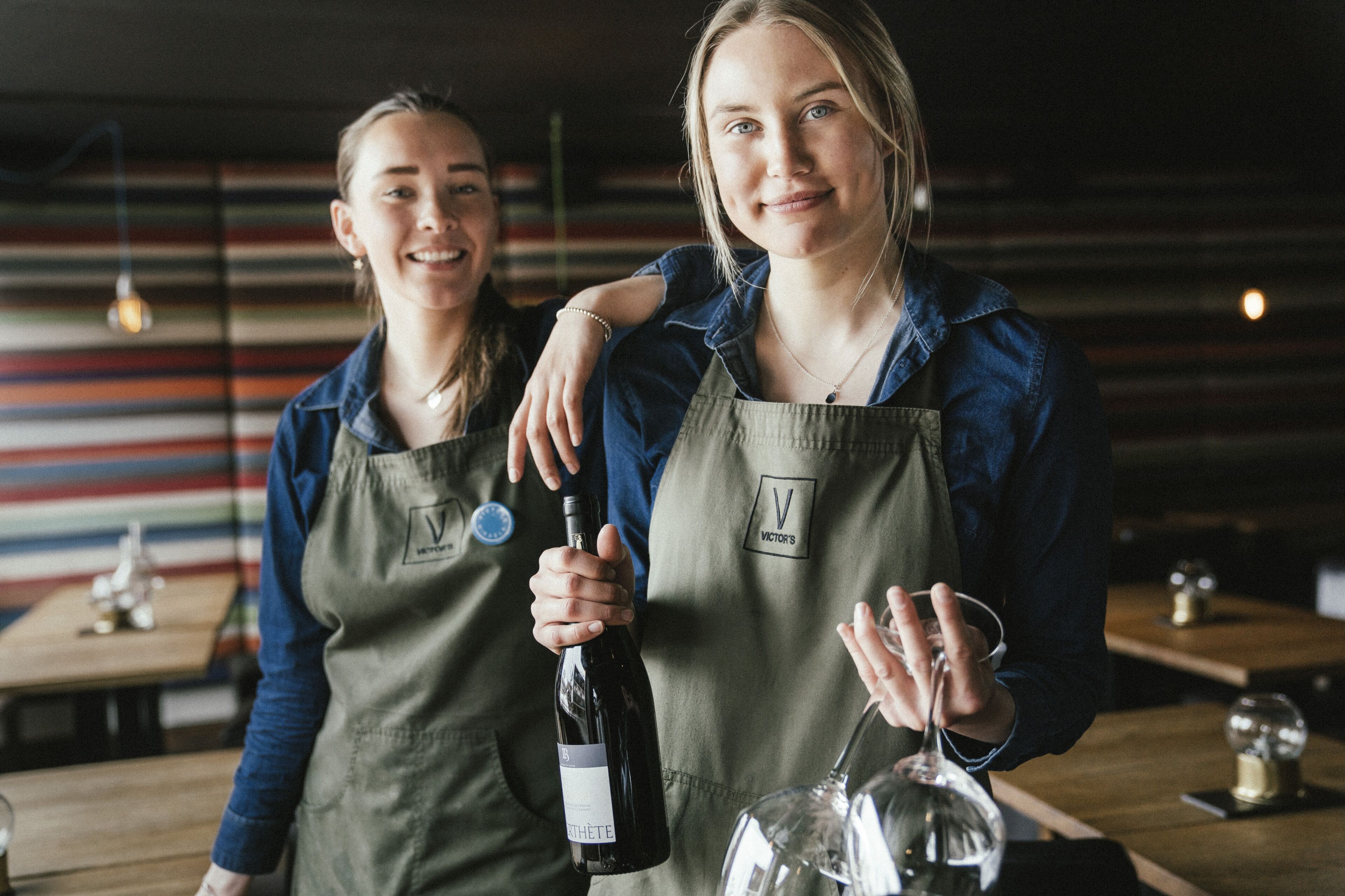 Dos personas con delantales, una botella de vino y una copa, en un ambiente con paredes a rayas y mesas de madera.