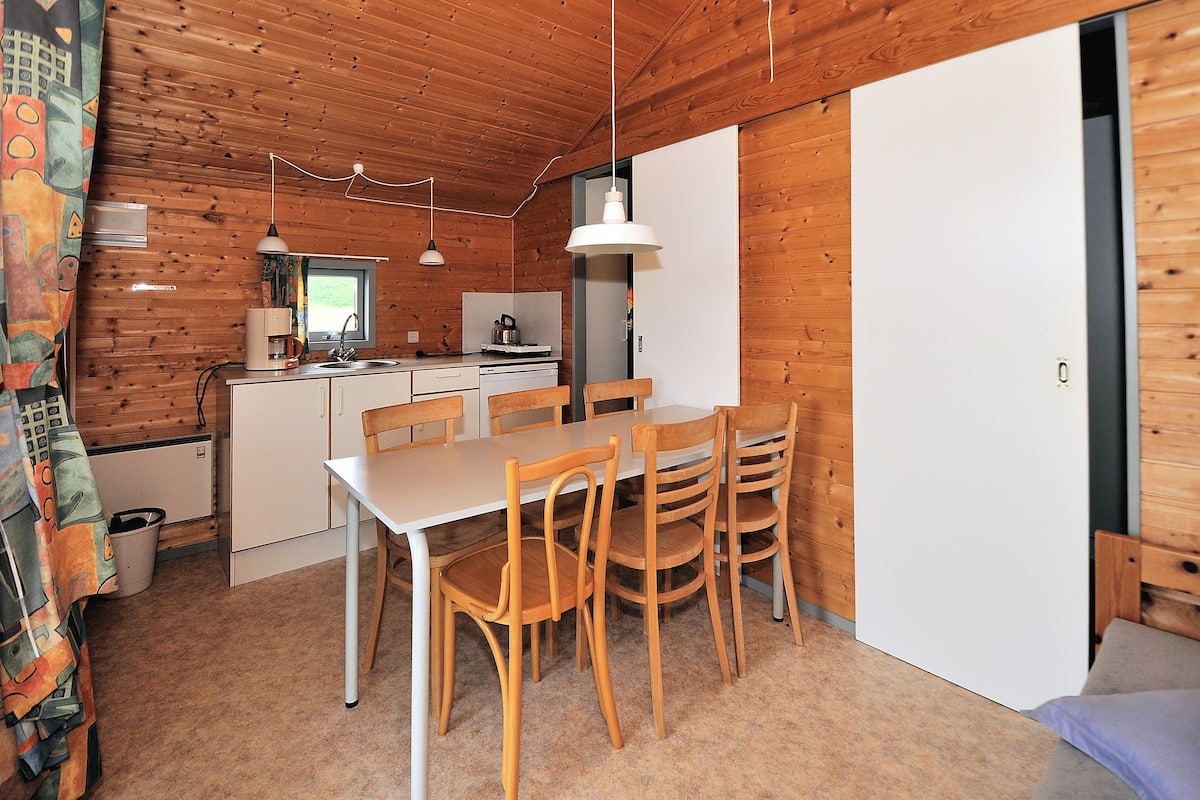A compact kitchen with wooden walls, a dining table with chairs, and a refrigerator.