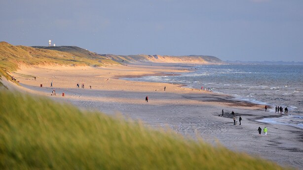 Une plage où l’on voit des gens se promener et se tenir debout, un phare en arrière-plan et des dunes herbeuses.