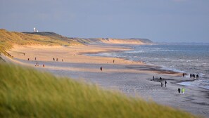 Een strand met mensen die lopen en staan, een vuurtoren in de verte en grasduinen.