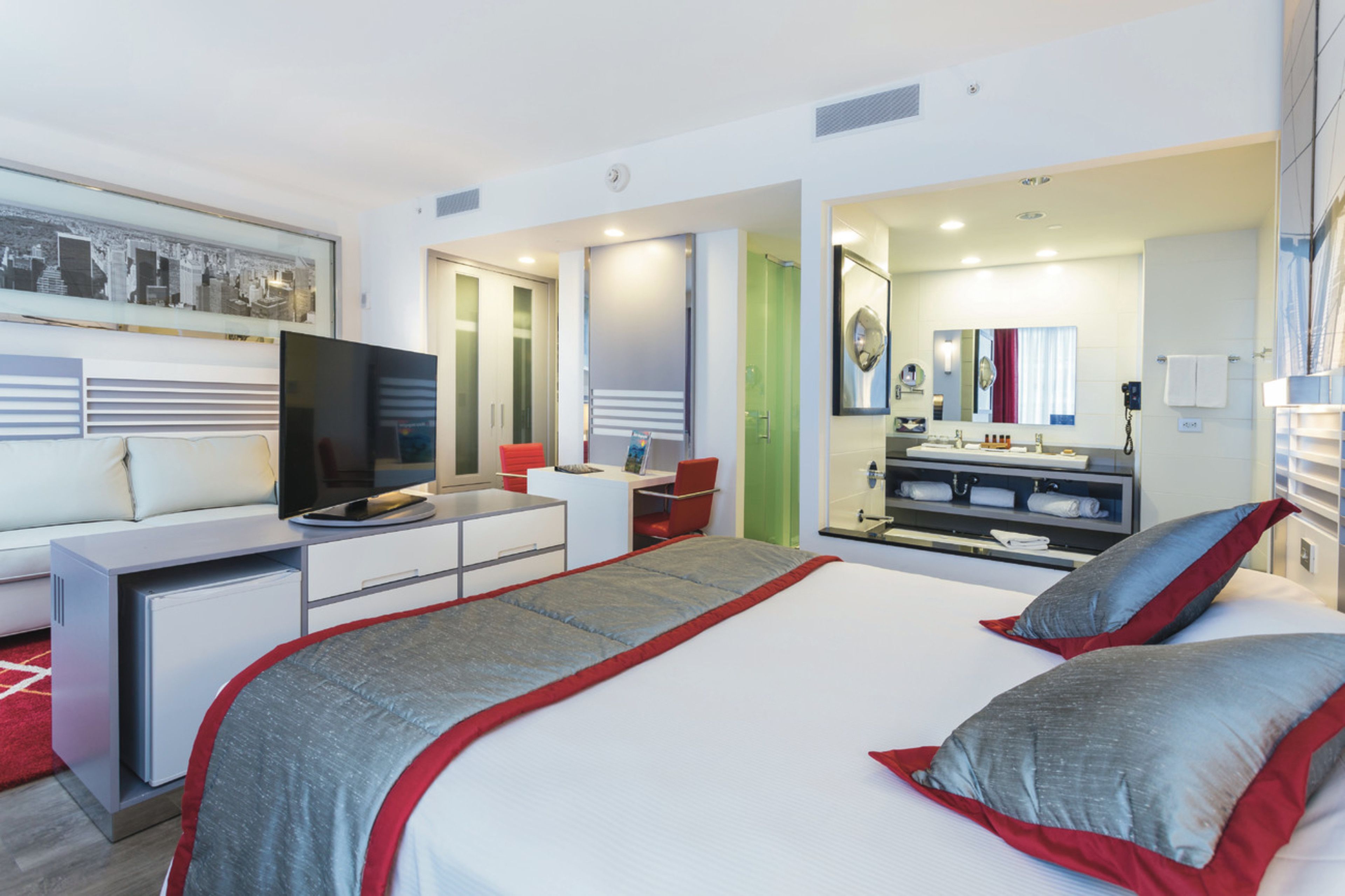 A modern hotel room with a bed, a desk, a television, a mirror, and a bathroom.