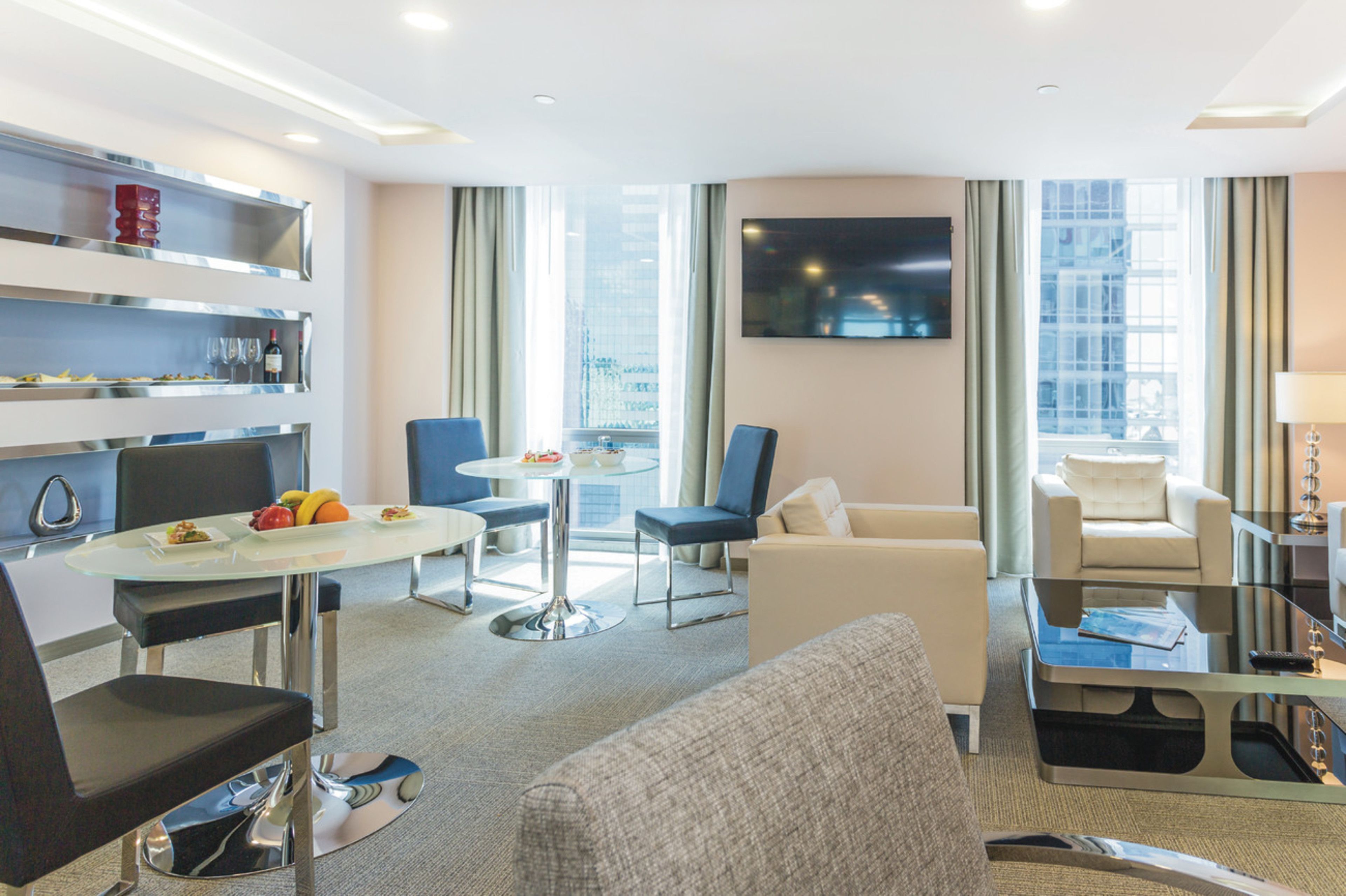 A modern hotel room with a glass dining table, two sofas, a TV mounted on the wall, and a city view through large windows.