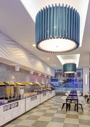 A modern hotel lobby with a checkered floor, a buffet area with various food items, and a unique blue cylindrical light fixture. - Hotel Riu Plaza New York Times Square (New York)