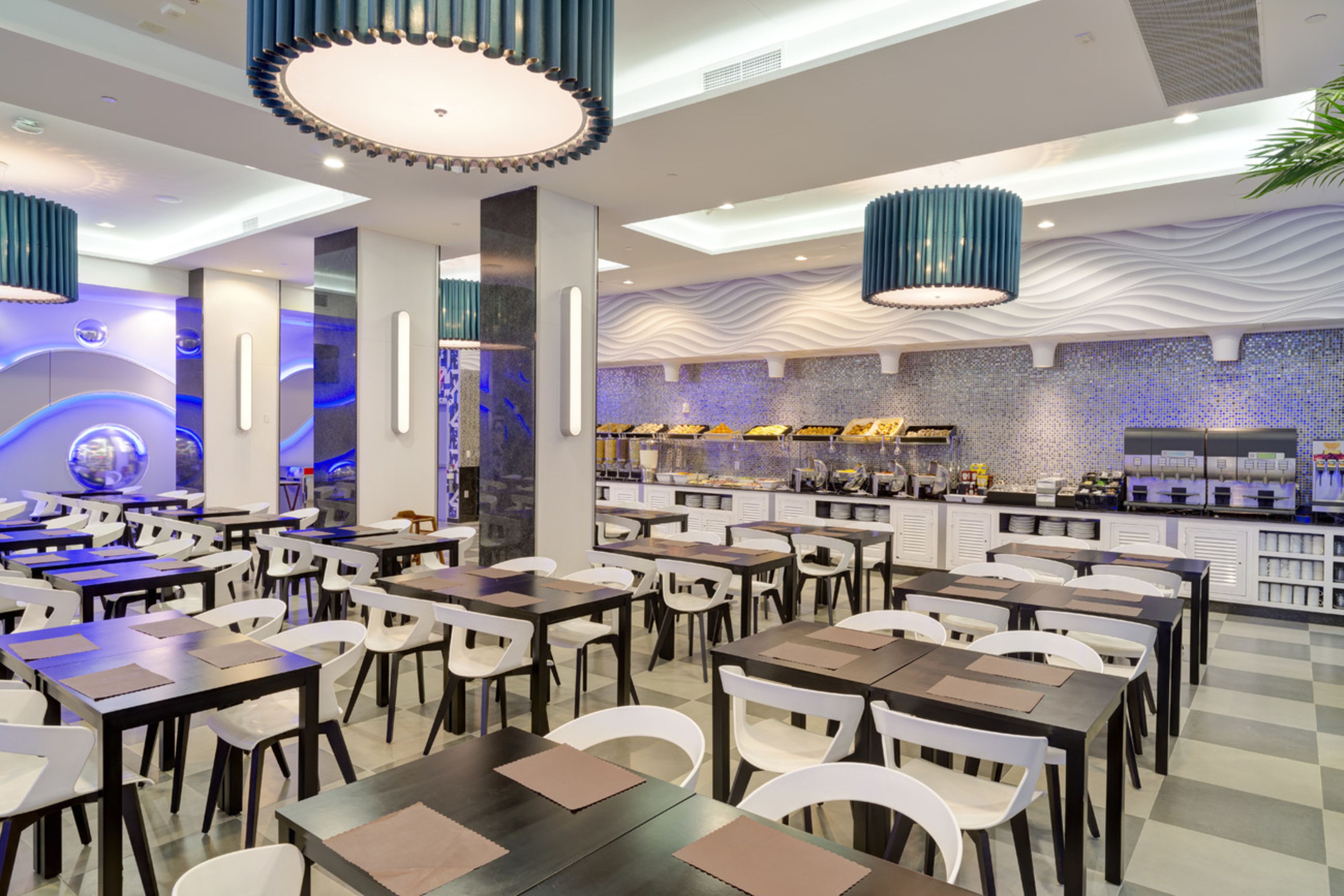 En moderne restaurant med flere spiseborde og stole, et centralt barområde og dekorativ belysning.