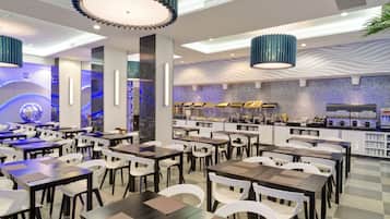 En moderne restaurant med flere spiseborde og stole, et centralt barområde og dekorativ belysning.