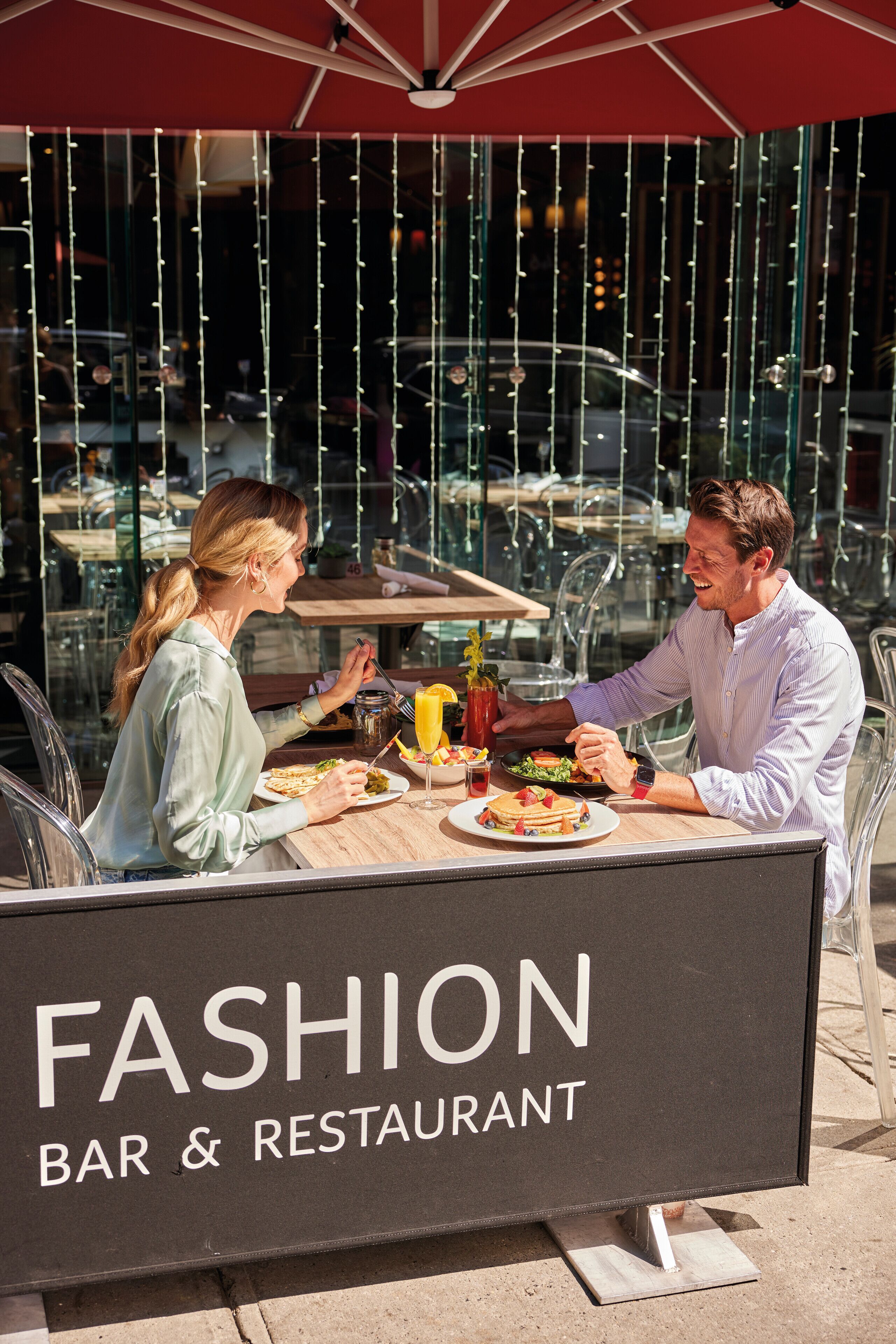 Ein Essbereich im Freien mit einem Tisch unter einem roten Sonnenschirm, einem Schild mit der Aufschrift 'FASHION BAR & RESTAURANT' und einer Glaskonstruktion im Hintergrund.