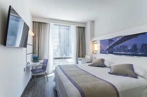A hotel room with a bed, a desk, a chair, a TV, and a large window with a cityscape view.