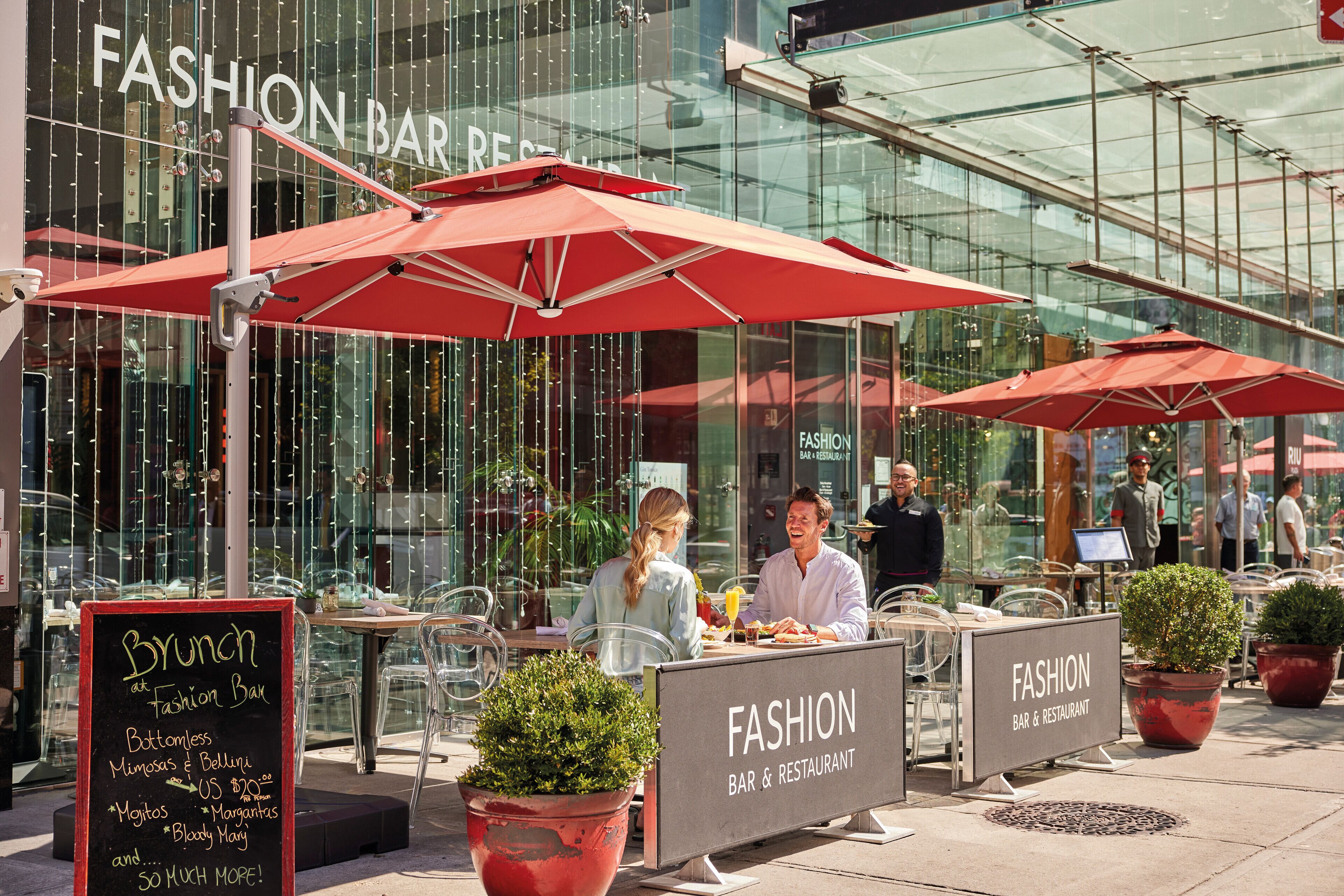 An outdoor dining area with red umbrellas, a sign for 'Fashion Bar & Restaurant', and a menu board listing brunch items.