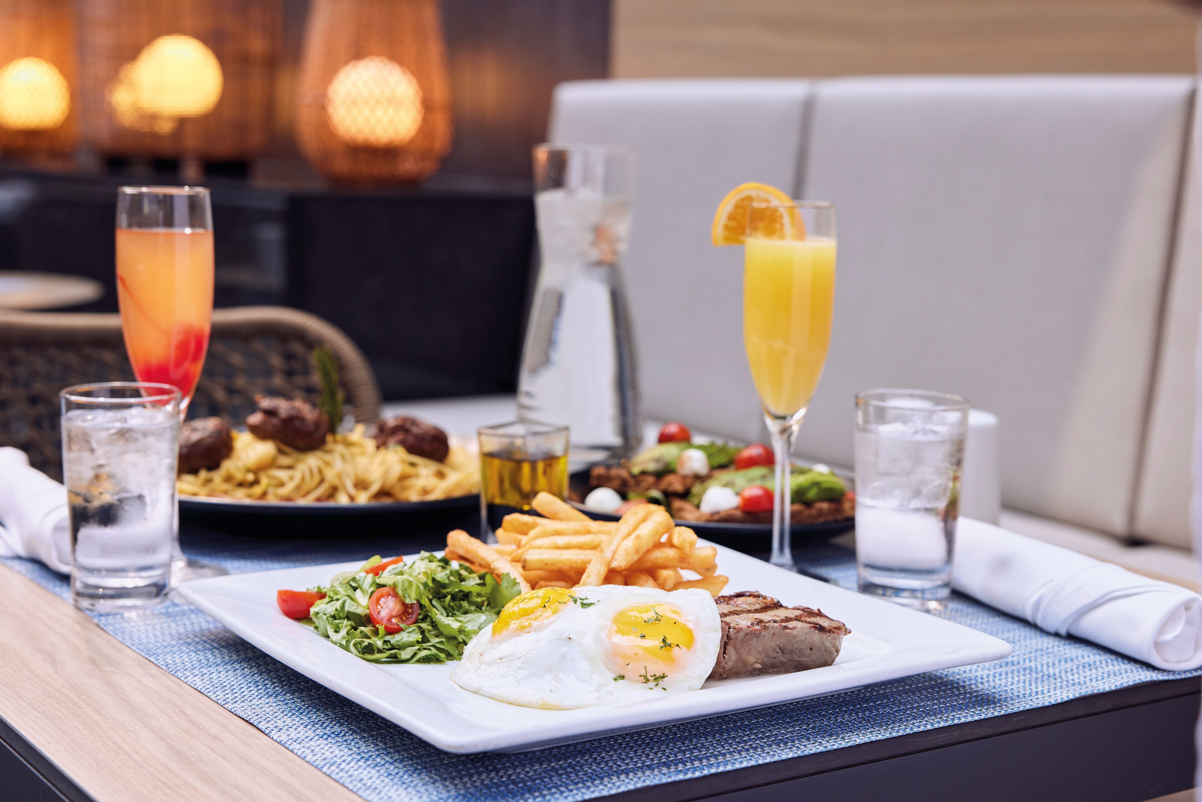 Una mesa de comedor con varios platos, incluyendo un bistec, un huevo frito, papas fritas, ensalada y un vaso de jugo de naranja.