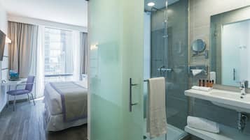 Quarto de hotel moderno com cama, escrivaninha, cadeira, box de chuveiro de vidro e banheiro com pia e espelho.