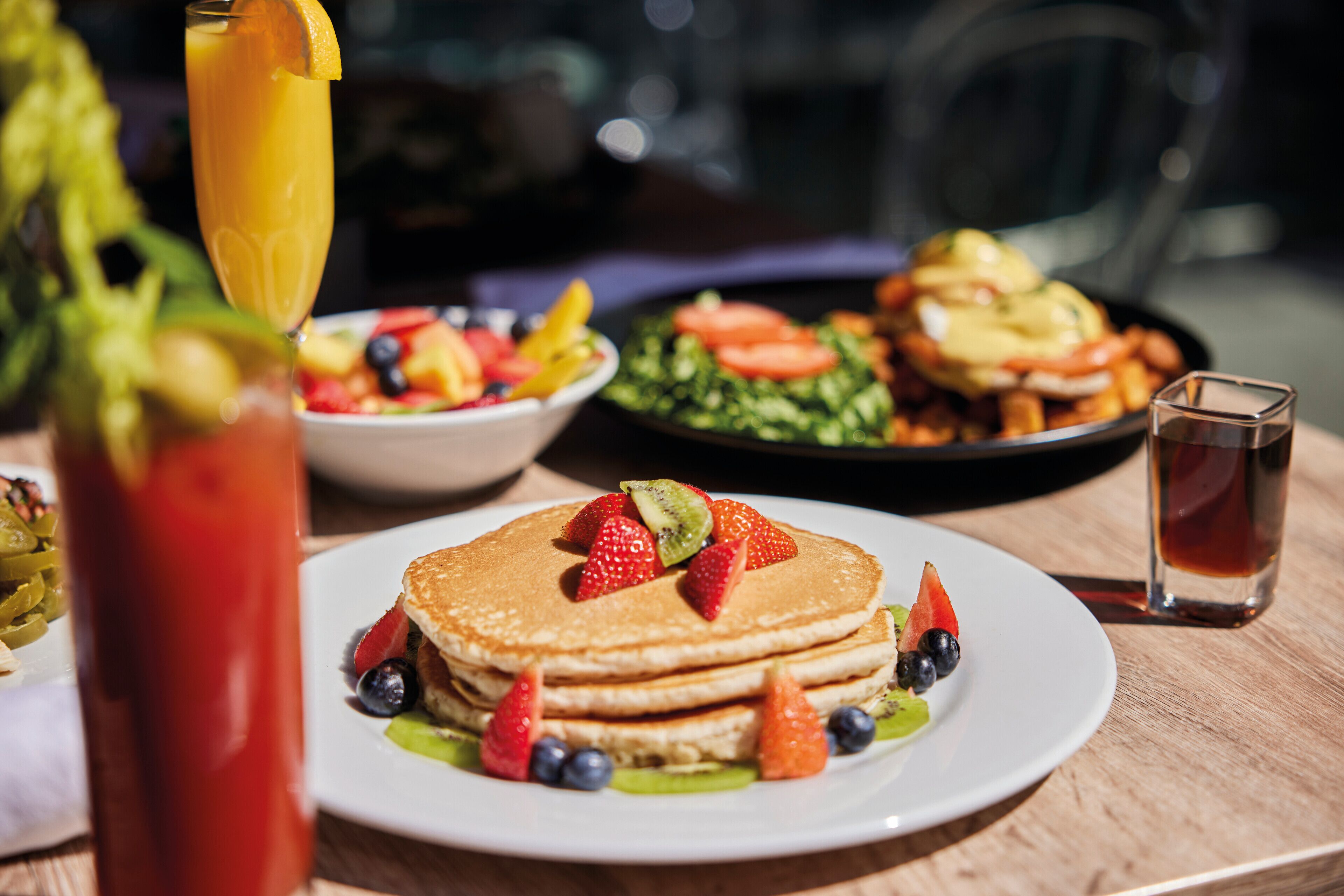 En frukostuppläggning med pannkakor dekorerade med jordgubbar, blåbär och kiwi, tillsammans med en sallad och ett glas juice.