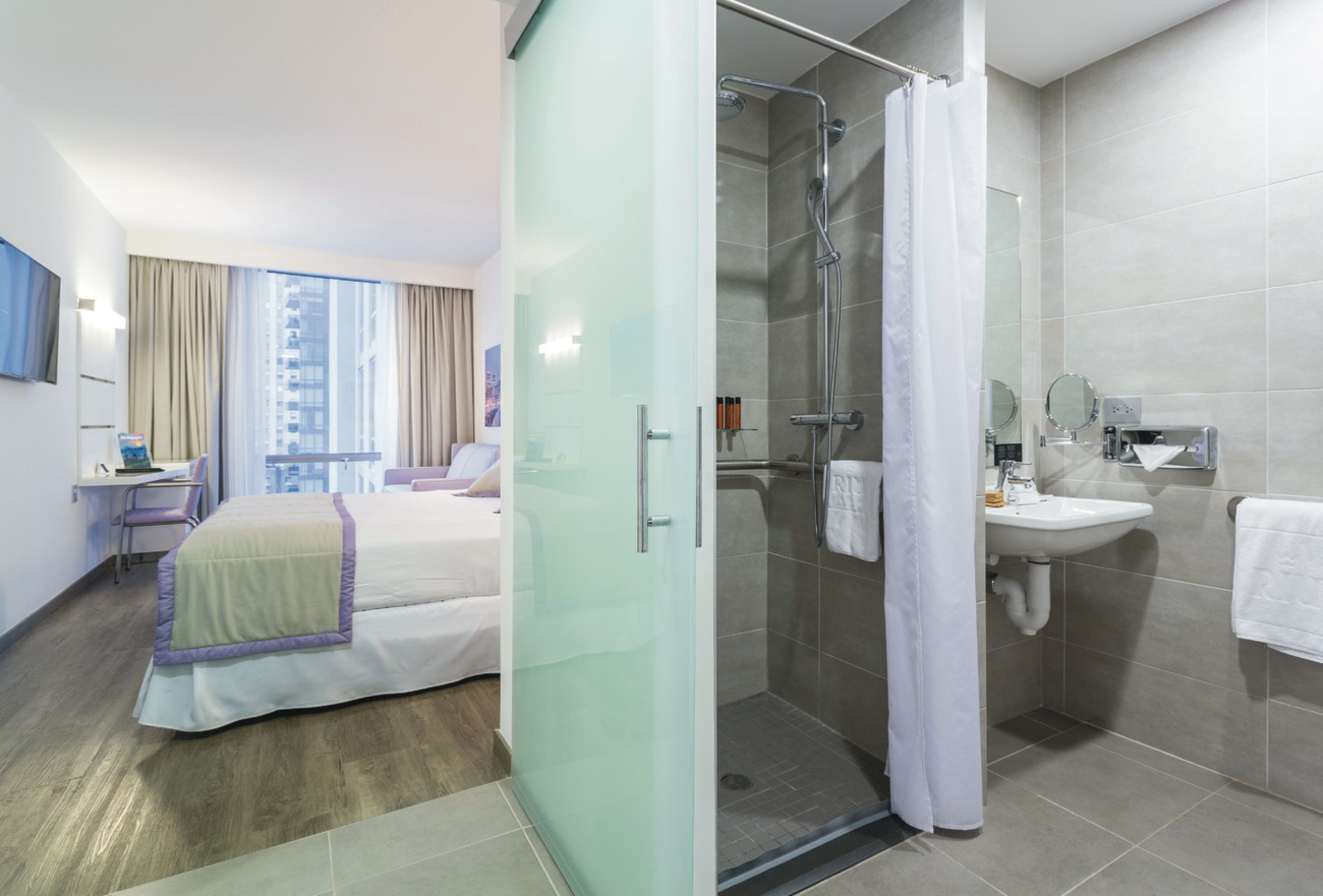 A hotel room with a bed, a desk with a laptop, a shower, and a bathroom with a sink and mirror.