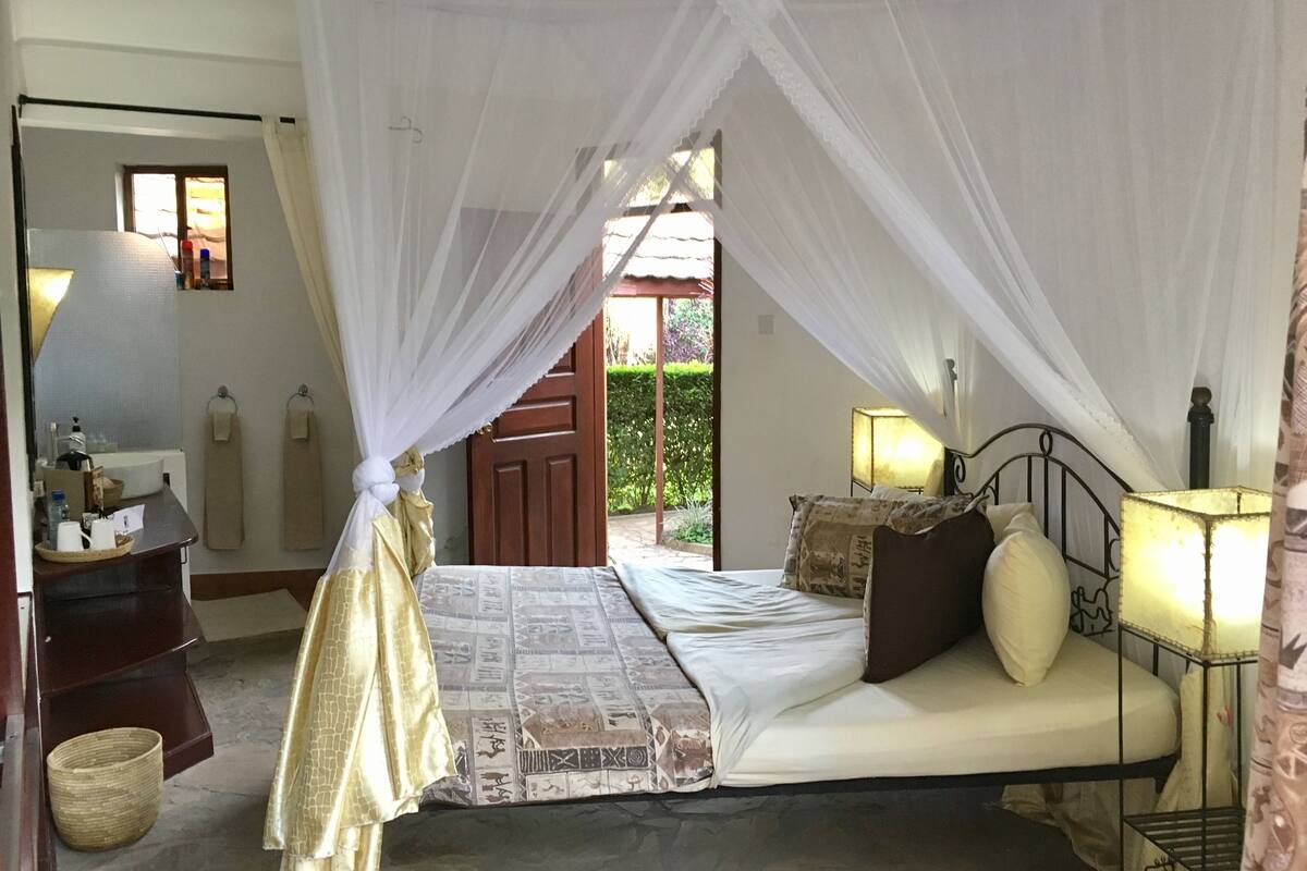 A bedroom with a bed under a canopy, a bedside table with a lamp, and a view of the outside.