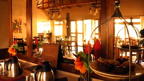 A cozy dining area with wooden beams, a large mirror, and a table set with a coffee pot, flowers, and a plate of muffins. - Ahadi Lodge (Arusha)