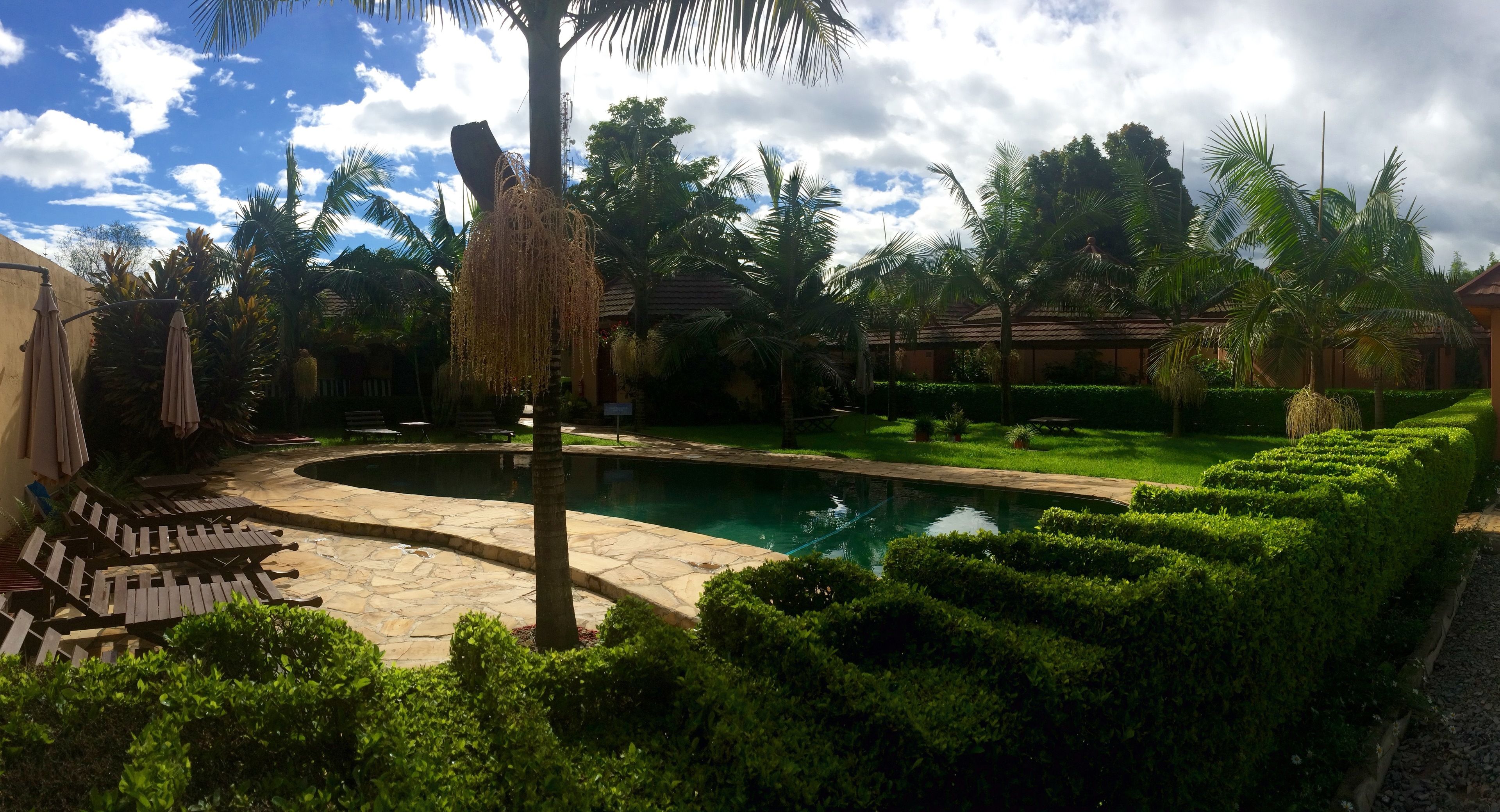 A pool area with stone flooring, palm trees, and neatly trimmed hedges.