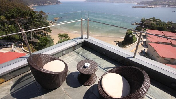 A balcony with wicker furniture and a clear view of the sea and coastline. - Apartamentos Las Sirenas (Viveiro)