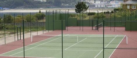 A tennis court with a green net and a red surface, surrounded by a green fence, with a beach and houses in the background.