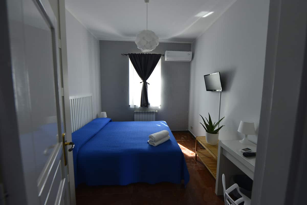Un dormitorio con cama, escritorio, silla, planta en maceta, aire acondicionado y ventana con cortinas.