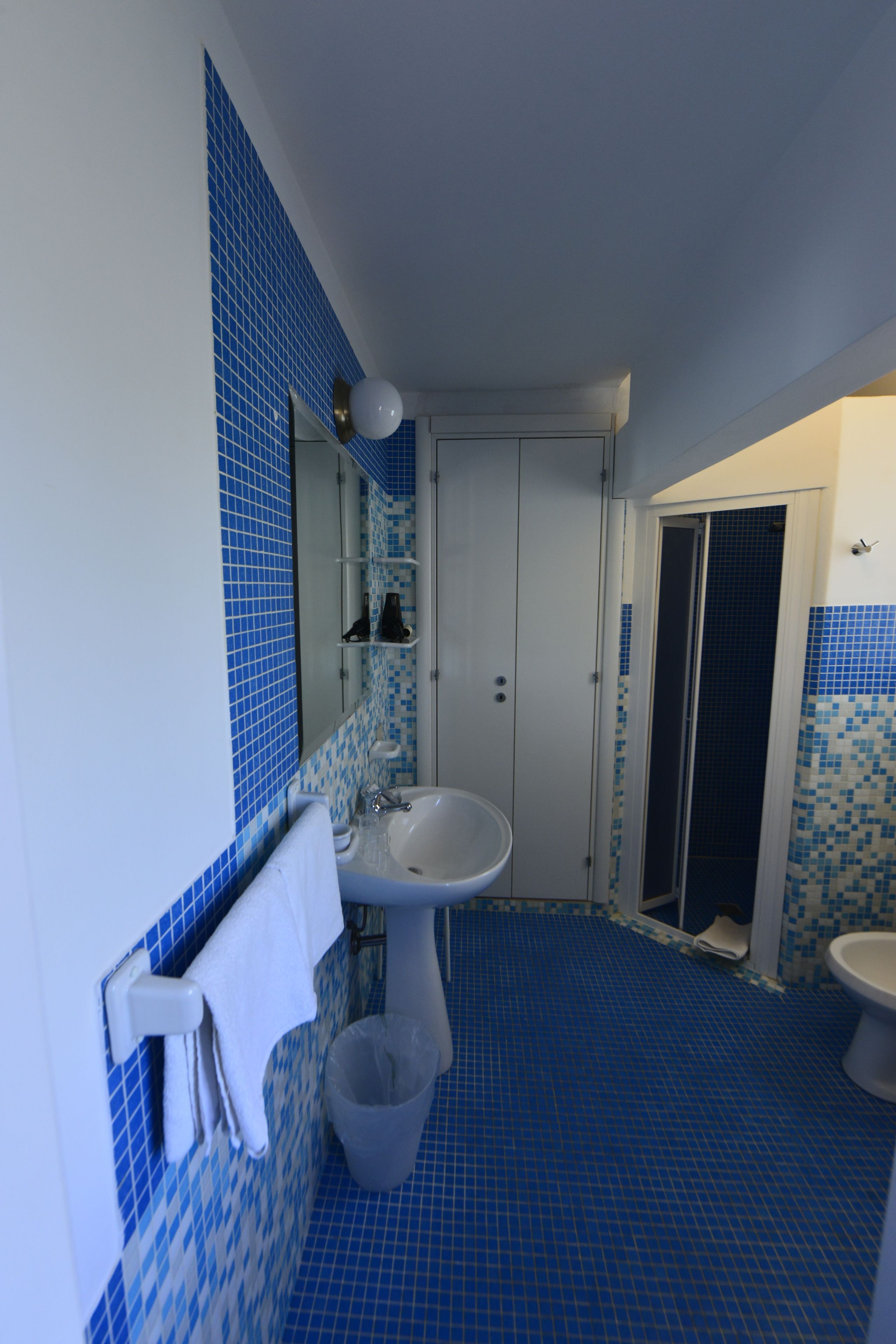 Un baño con lavamanos, espejo y retrete. El suelo y las paredes están cubiertos de azulejos azules.