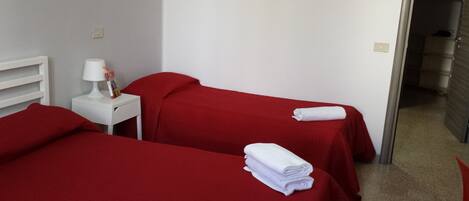 Habitación de hotel con dos camas, cada una con una colcha roja y una toalla blanca al final. Se ve un mesita de noche blanca con una lámpara y un objeto pequeño encima.