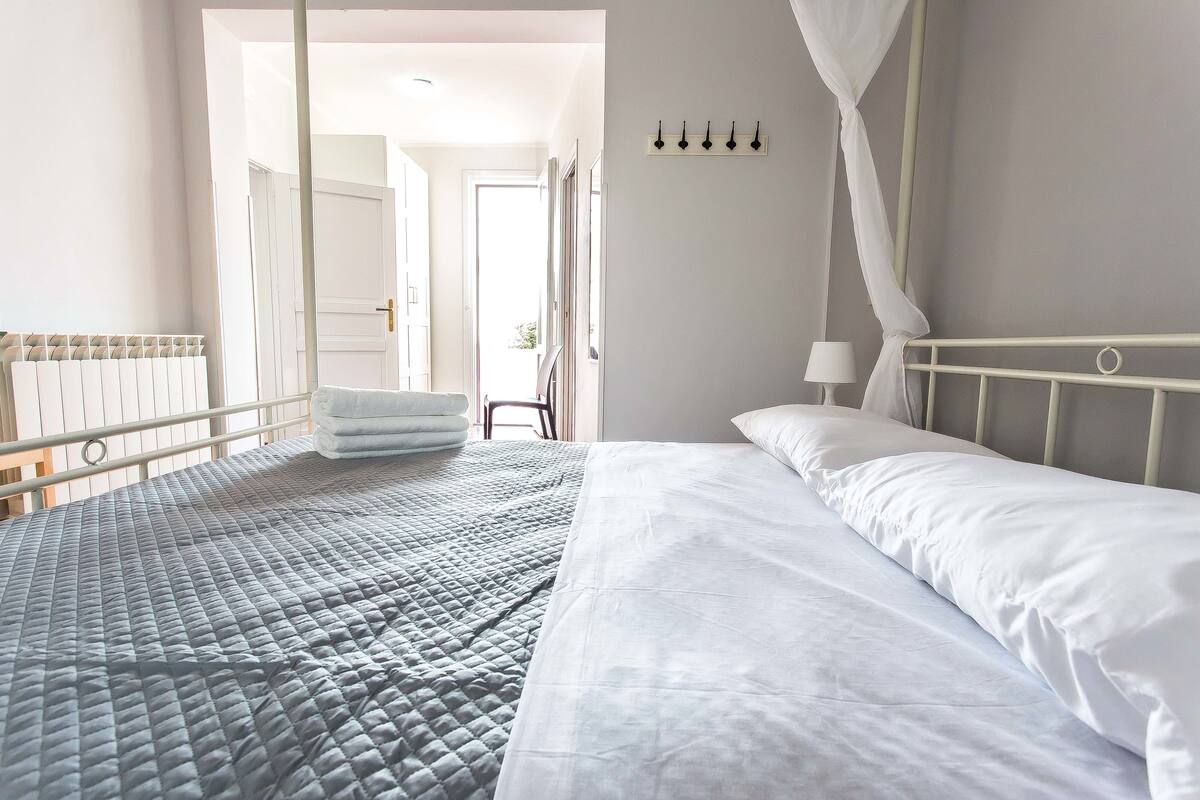 Una cama bien hecha con colcha gris y ropa de cama blanca, un radiador blanco y un dosel blanco.