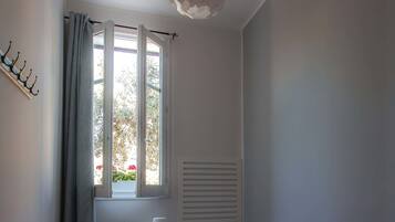Una habitación pequeña y bien iluminada con una cama, una mesita de noche con lámpara, una silla, una ventana con cortinas y una lámpara colgante.