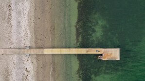An aerial view of a wooden pier extending into a body of water, with a small structure at the end.