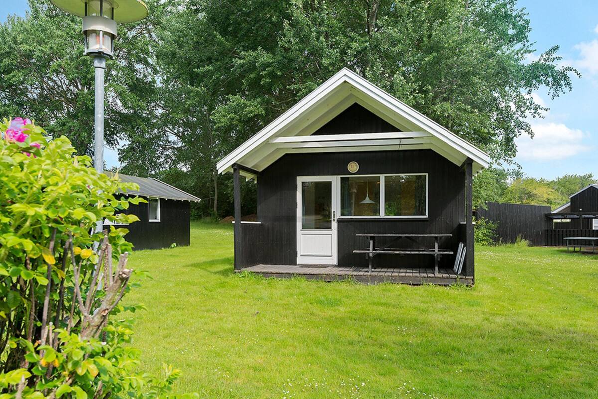 A small, single-story building with a white door and windows, surrounded by a grassy area and trees.