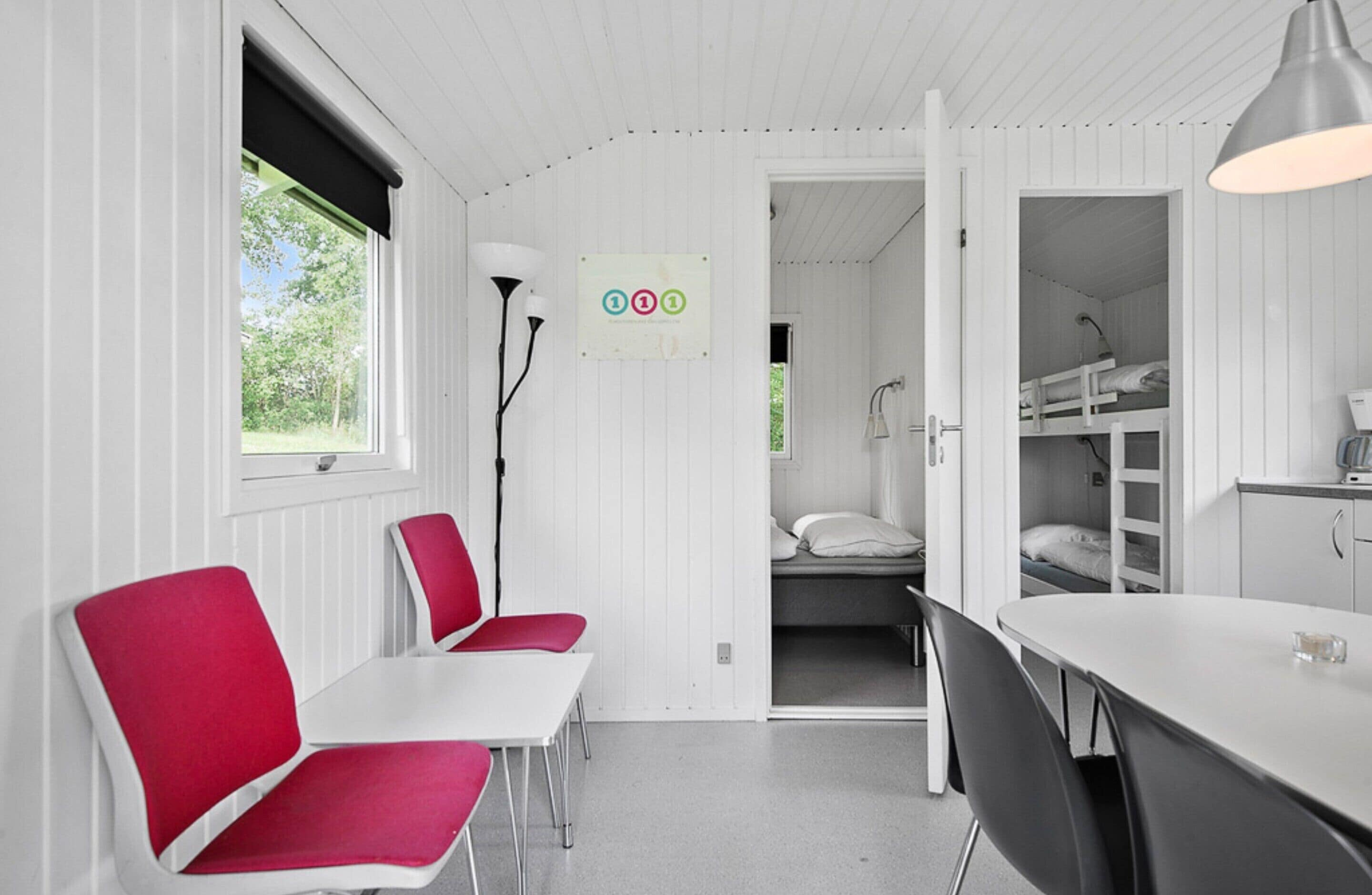 Een compacte kamer met witte wanden en een wit plafond, met een eettafel met rode stoelen, een witte tafel en een bed met een witte hoofdeinde.