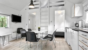 A modern kitchen with white cabinetry, a dining area with black chairs, a TV mounted on the wall, and a window providing natural light. - First Camp Ajstrup Strand (Malling)