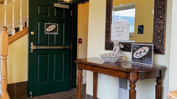 Un hall de hotel con una puerta verde, una escalera de madera, un espejo con marco decorativo e información sobre estacionamiento en una mesa.