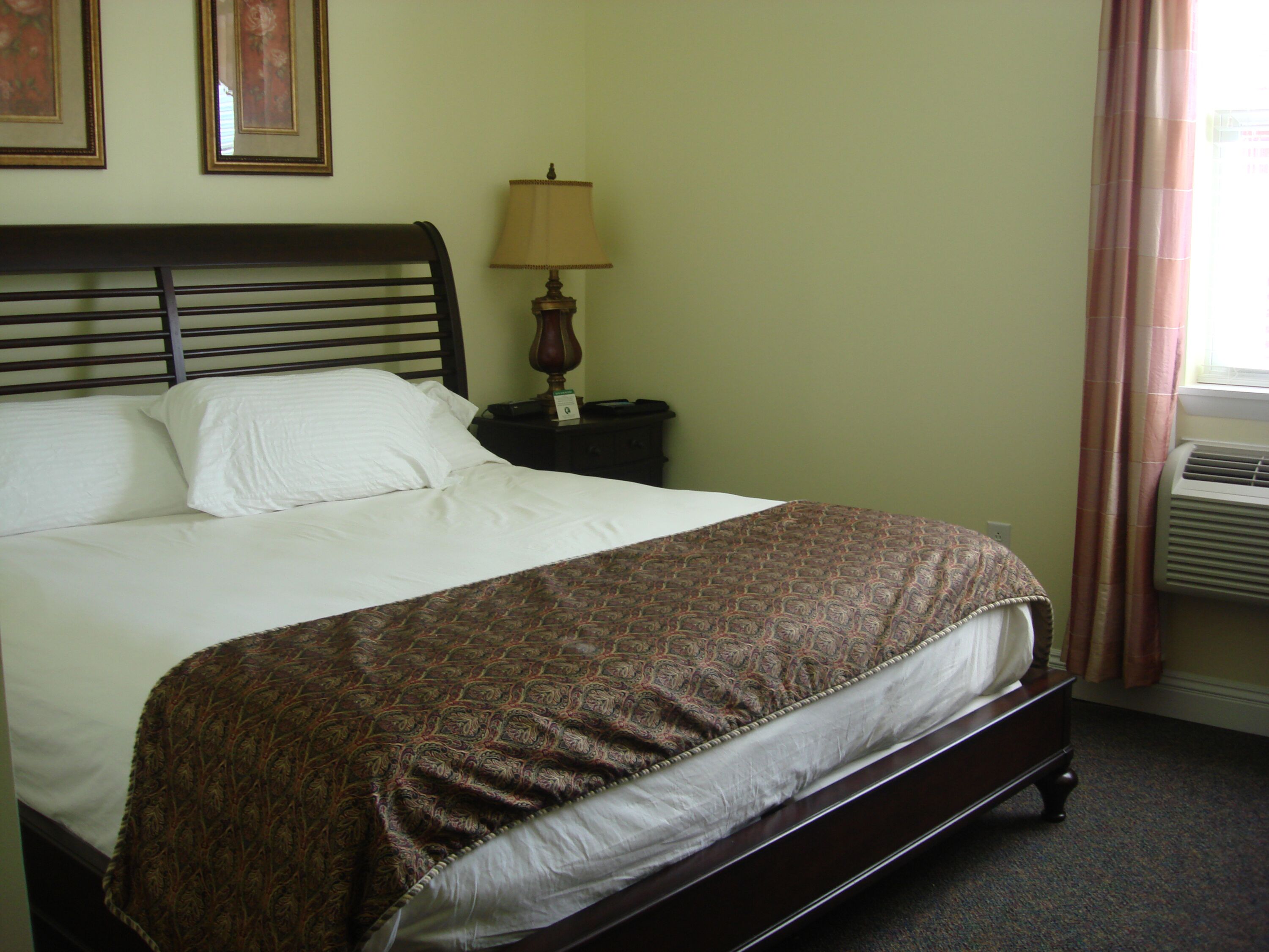 A neatly made bed with a patterned bedspread, a wooden headboard, a bedside table with a lamp, and a wall-mounted air conditioner.