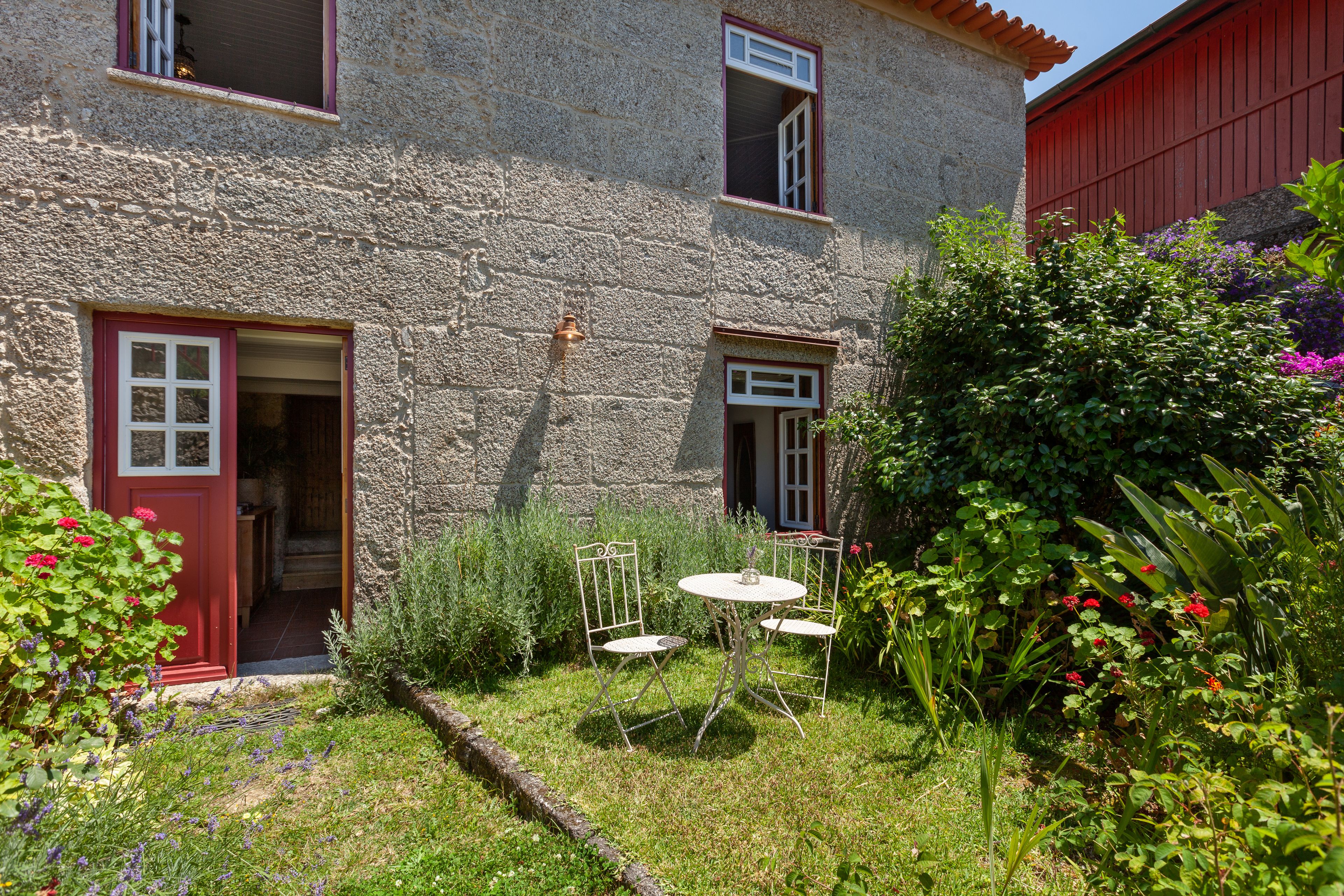 A stone building with a red door and windows, a small garden with a table and chairs, and various plants and flowers.