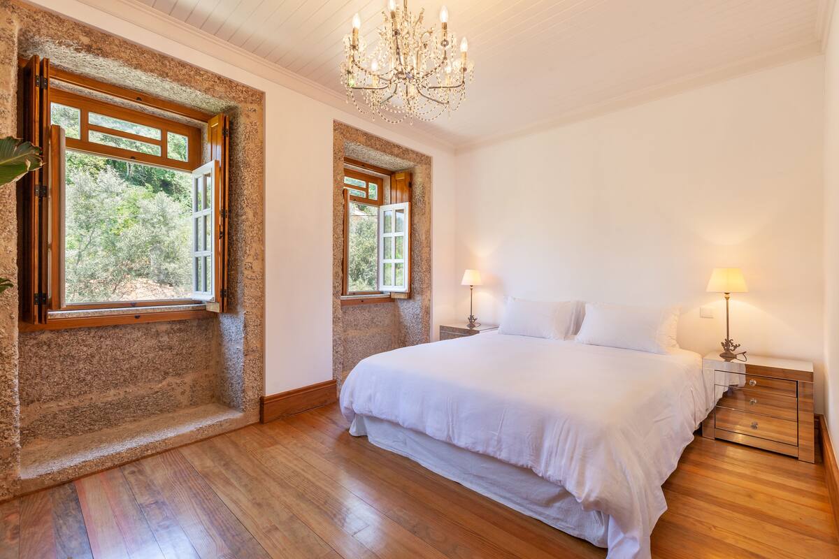 Une chambre avec un grand lit, deux fenêtres, un lustre et un plancher en bois.