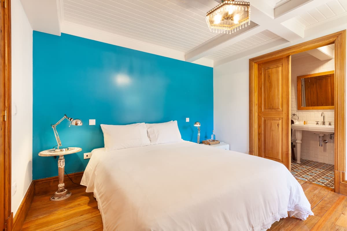 Une chambre avec un grand lit, une table de chevet avec une lampe, un mur d’accent bleu et une porte en bois donnant sur une salle de bain.