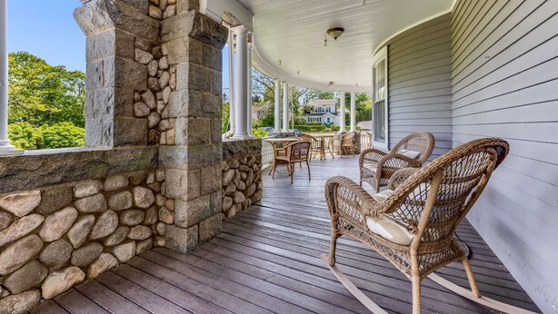 A covered porch with stone walls, wicker furniture, and a view of a house in the background. - Castle Manor Inn (Gloucester)
