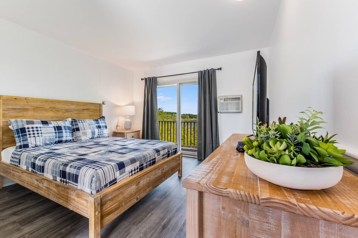 Un dormitorio con una cama de madera, una mesita de noche, un ventanal con cortinas, un televisor y una planta en maceta.
