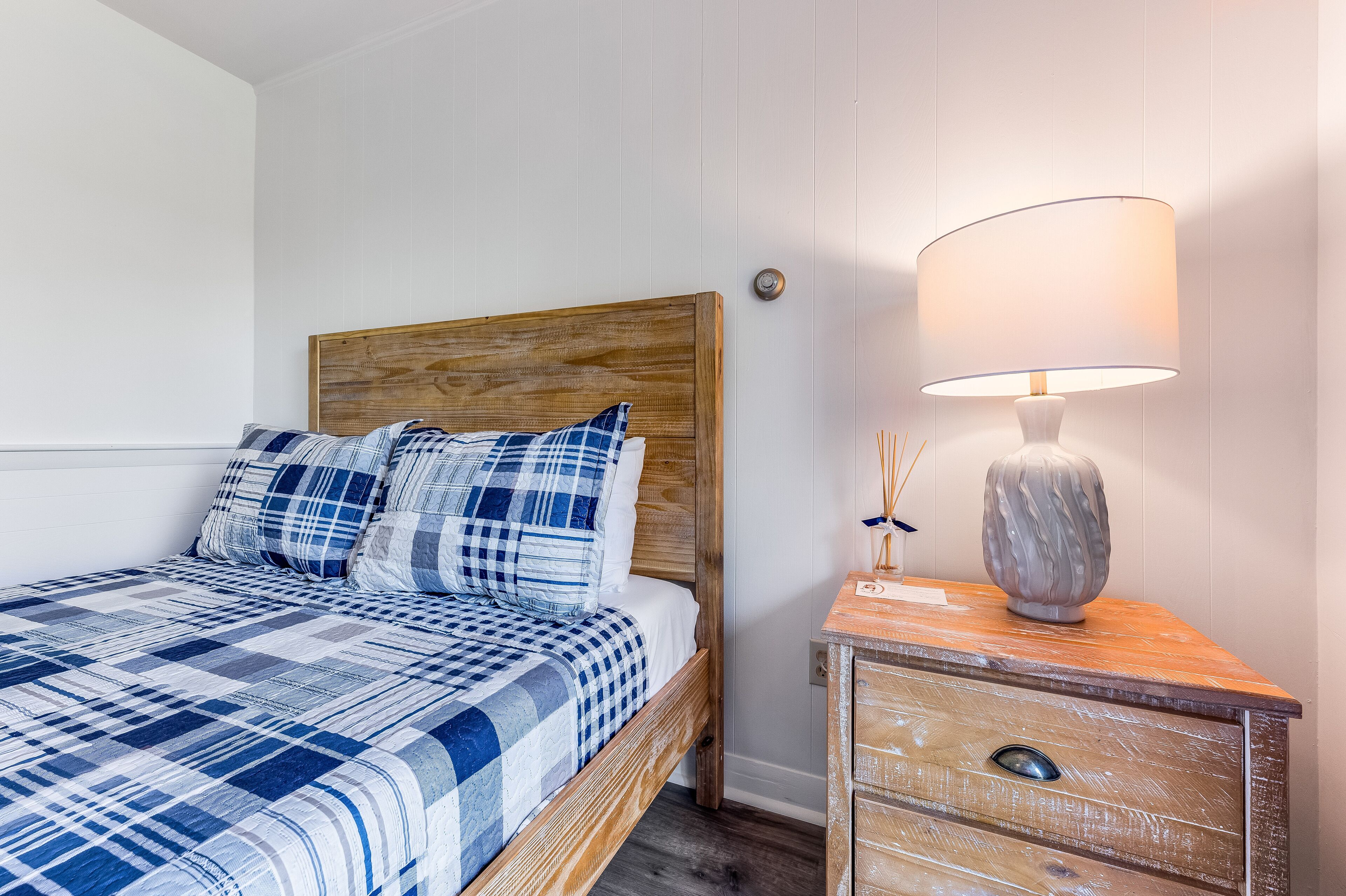 Una cama con colcha azul y blanca a cuadros, cabecera de madera y mesita de noche con lámpara y objetos decorativos.