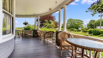 A covered outdoor deck with wicker furniture and a glass table, overlooking a well-maintained garden and a road.