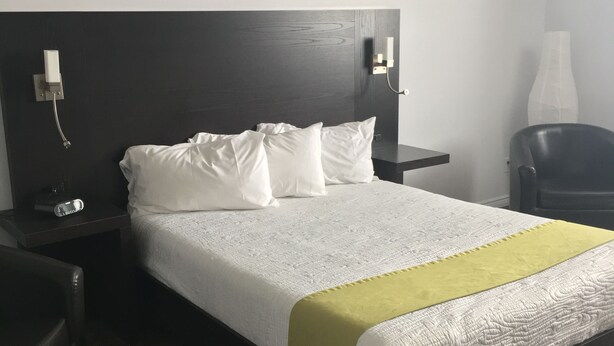 A neatly made bed with white bedding and a green accent, a dark wooden headboard, a bedside table with a phone and a lamp, and a chair in a hotel room.