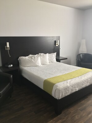A neatly made bed with white bedding and a green accent, a dark wooden headboard, a bedside table with a phone and a lamp, and a chair in a hotel room. - Hotel 1212 (Degelis)