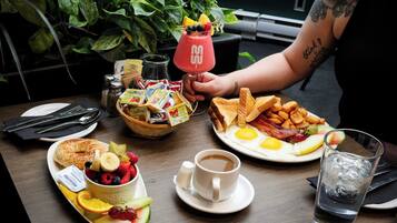 Mesa de café da manhã com diversos alimentos, incluindo torradas, ovos, bacon e frutas. Há uma xícara de café e um copo de água sobre a mesa.