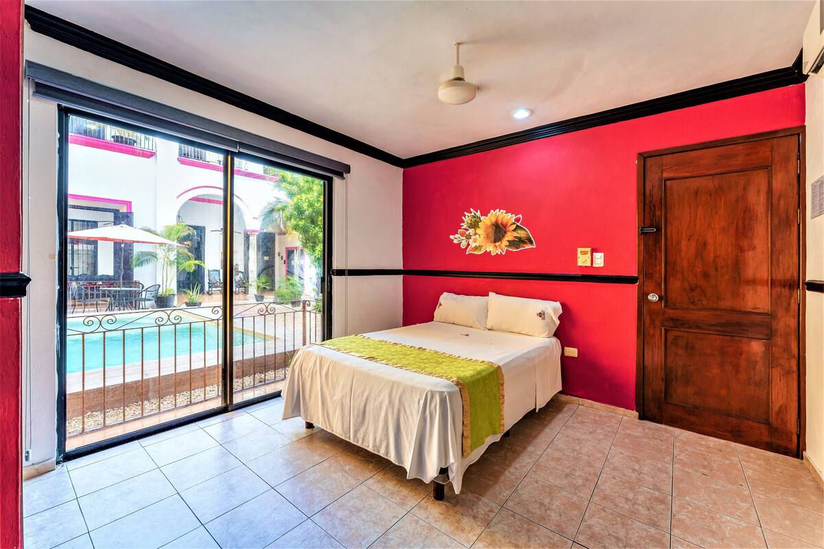 A bedroom with a bed, a red accent wall, a wooden door, and a sliding glass door leading to an outdoor pool area.
