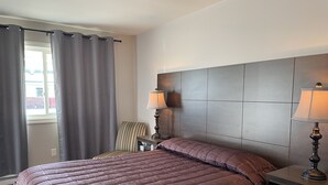 A hotel room with a large bed, a headboard, two bedside lamps, a chair, and a window with curtains. - Winsome Inn (St. Stephen)