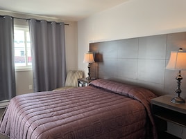Habitación de hotel con una cama grande, cabecera, dos lámparas de noche, una silla y una ventana con cortinas.