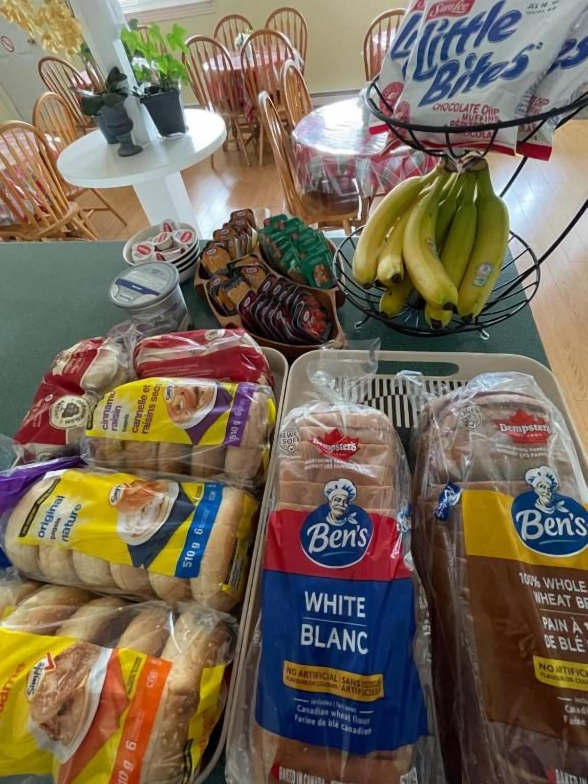 A dining area with a table set for a meal, including bread, bananas, and packaged snacks.