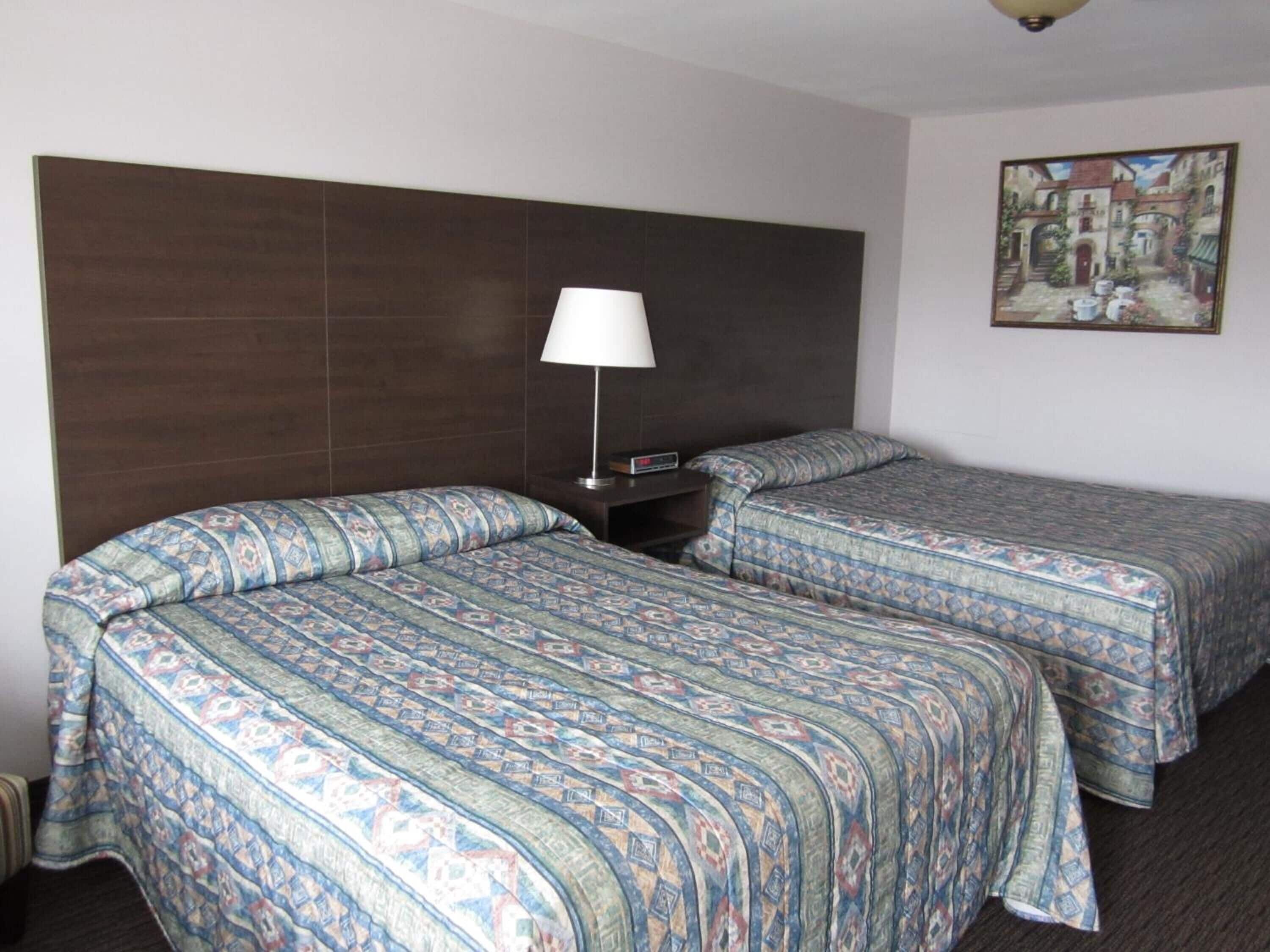 A hotel room with two beds, a nightstand, a lamp, and a painting on the wall.
