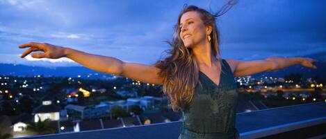 A woman standing on a balcony with arms outstretched, overlooking a cityscape at dusk.