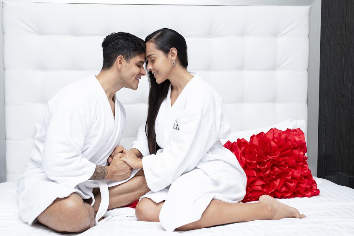 Una pareja sentada en una cama con cabecera blanca y un cojín decorativo rojo.